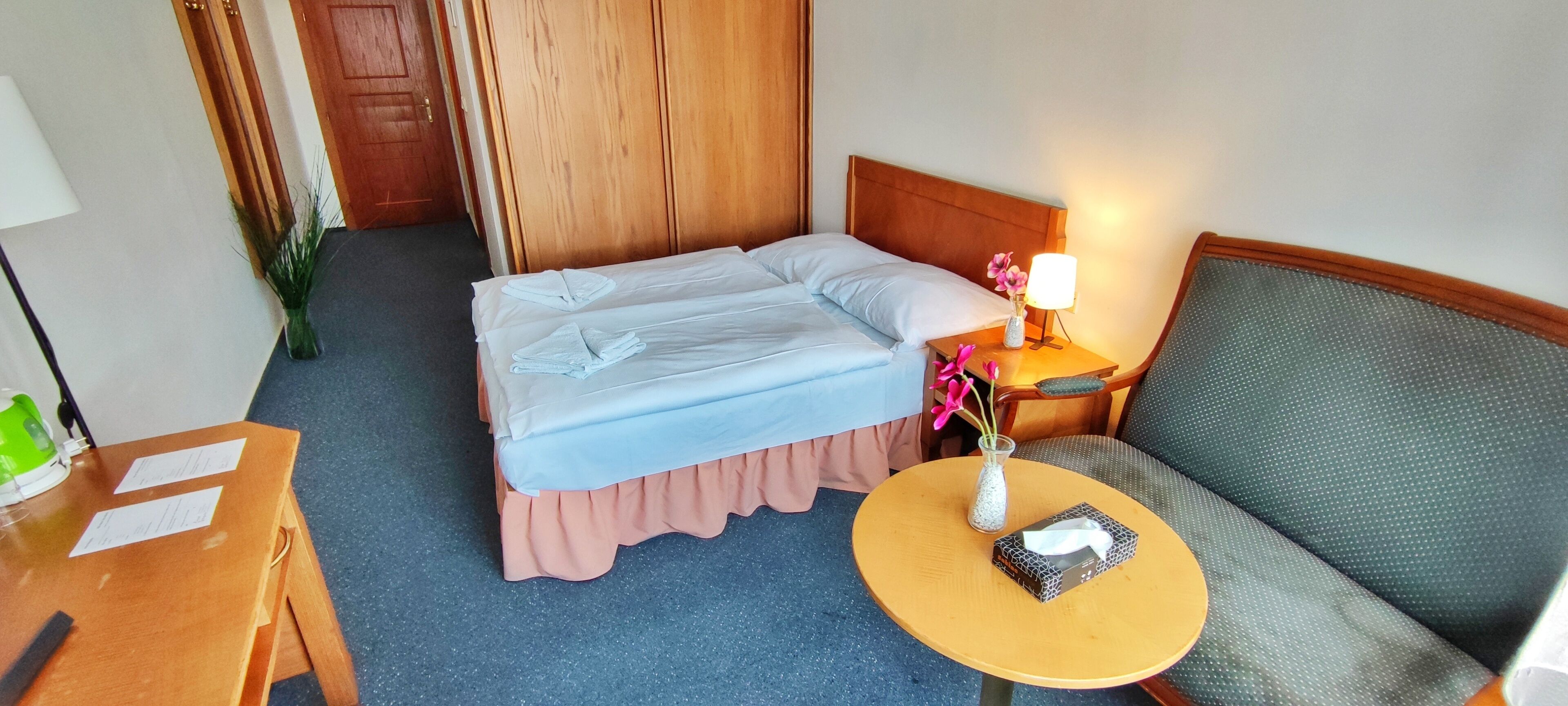 A hotel room with a bed, a small table with a lamp, a chair, and a vase with flowers.