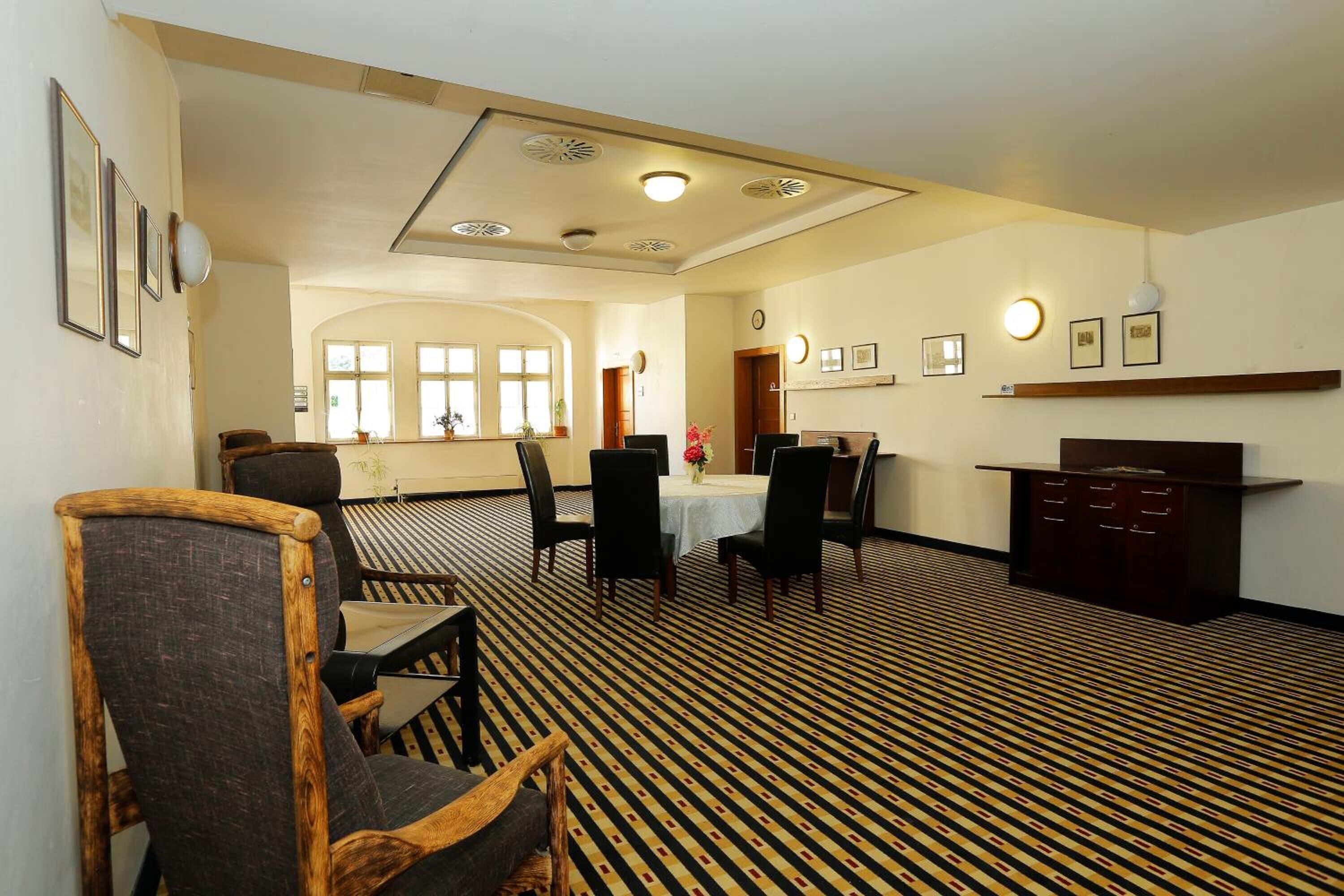 A dining area with a patterned carpet, wooden chairs, a table with a vase of flowers, and a wooden cabinet.