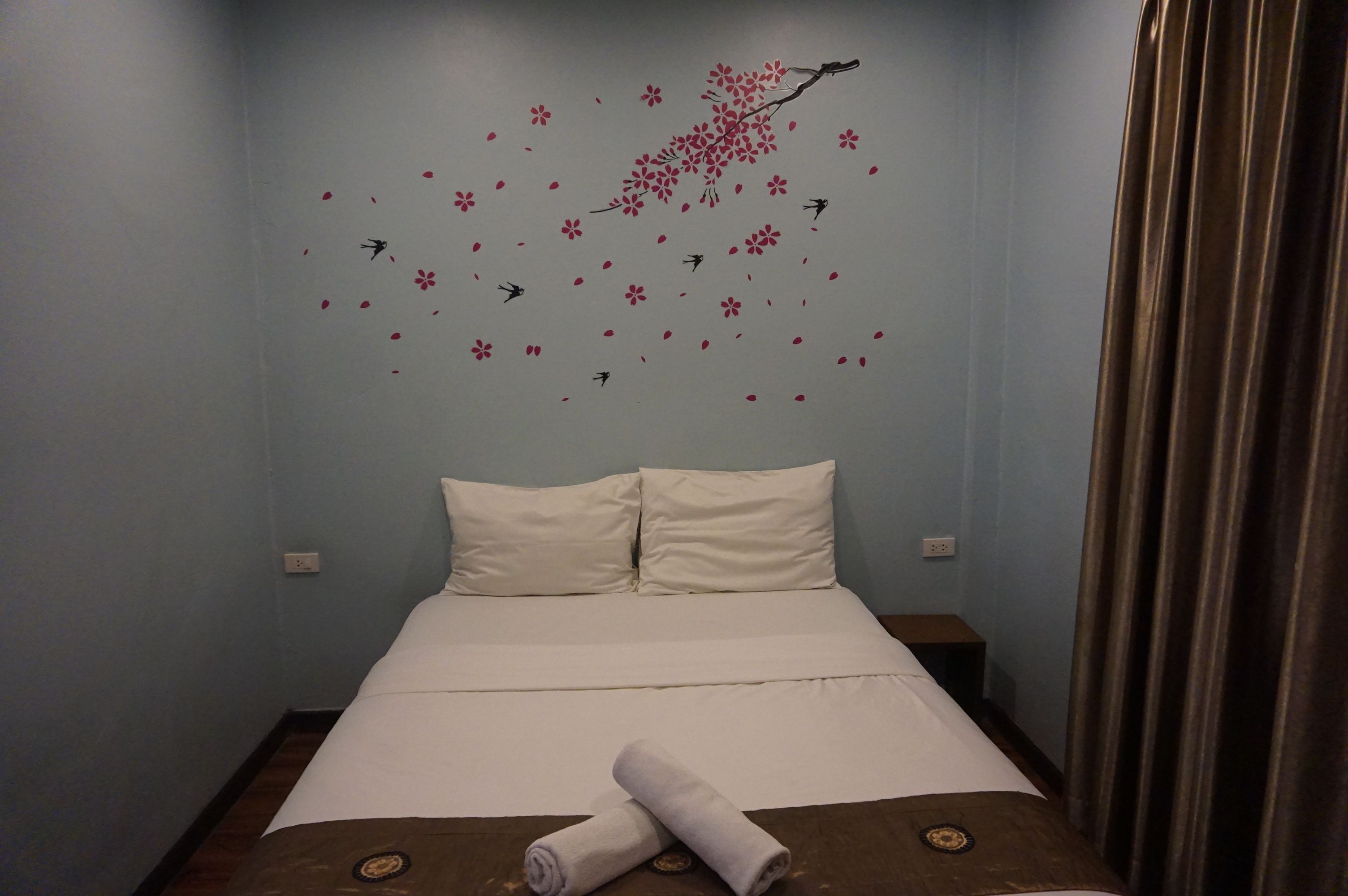 A neatly made bed with white linens, a pair of rolled towels, and a wall decoration of flowers and birds.
