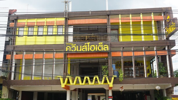 A multi-story building with a yellow and orange facade, a visible sign with Thai script, and a parking area in front. - Queens Hotel (Chum Phae)