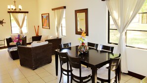 A dining area with a dark wooden table, six chairs, and a vase with flowers. There is a sofa and a chair in the background, and a painting on the wall. - Casa Dror Gran Pacifica Resort (Villa El Carmen)