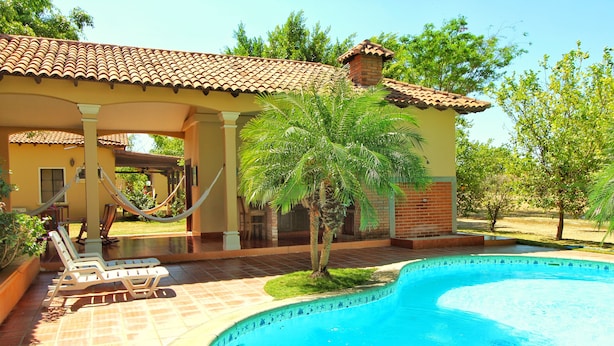 A swimming pool with a tiled deck, lounge chairs, and a palm tree. - Casa Dror Gran Pacifica Resort (Villa El Carmen)