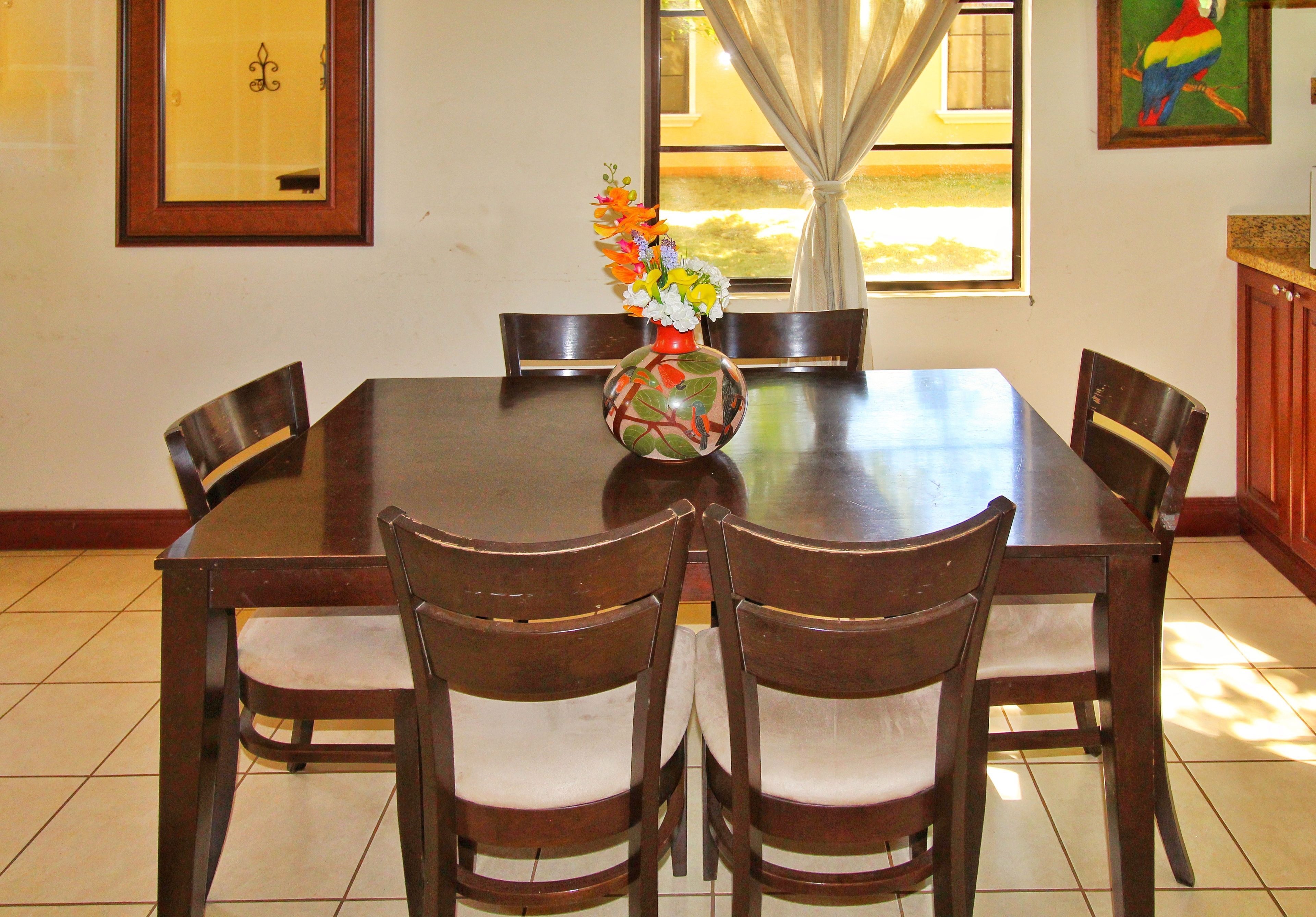 A dining table with four chairs, a vase with flowers, a window with curtains, and a framed picture on the wall.