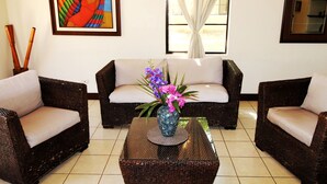 A room with two wicker sofas, a glass coffee table with a vase of flowers, a framed painting on the wall, and a window with curtains. - Casa Dror Gran Pacifica Resort (Villa El Carmen)