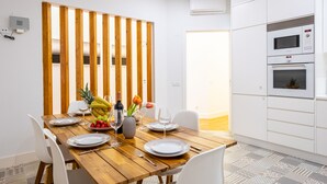 Een eetgedeelte met een houten tafel gedekt voor vier personen, witte stoelen en een keuken met witte kasten en apparatuur.