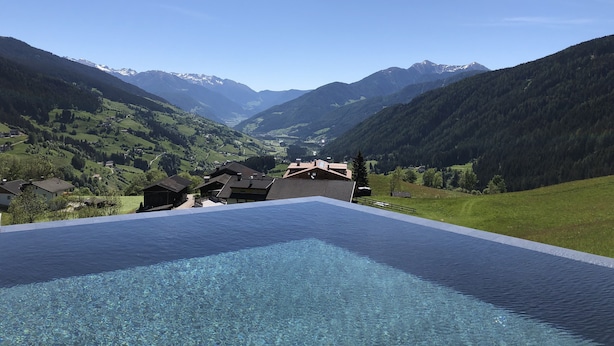 A pool with a mountain view, surrounded by houses and lush greenery. - Erlebnishotel Gassenhof (Racines)