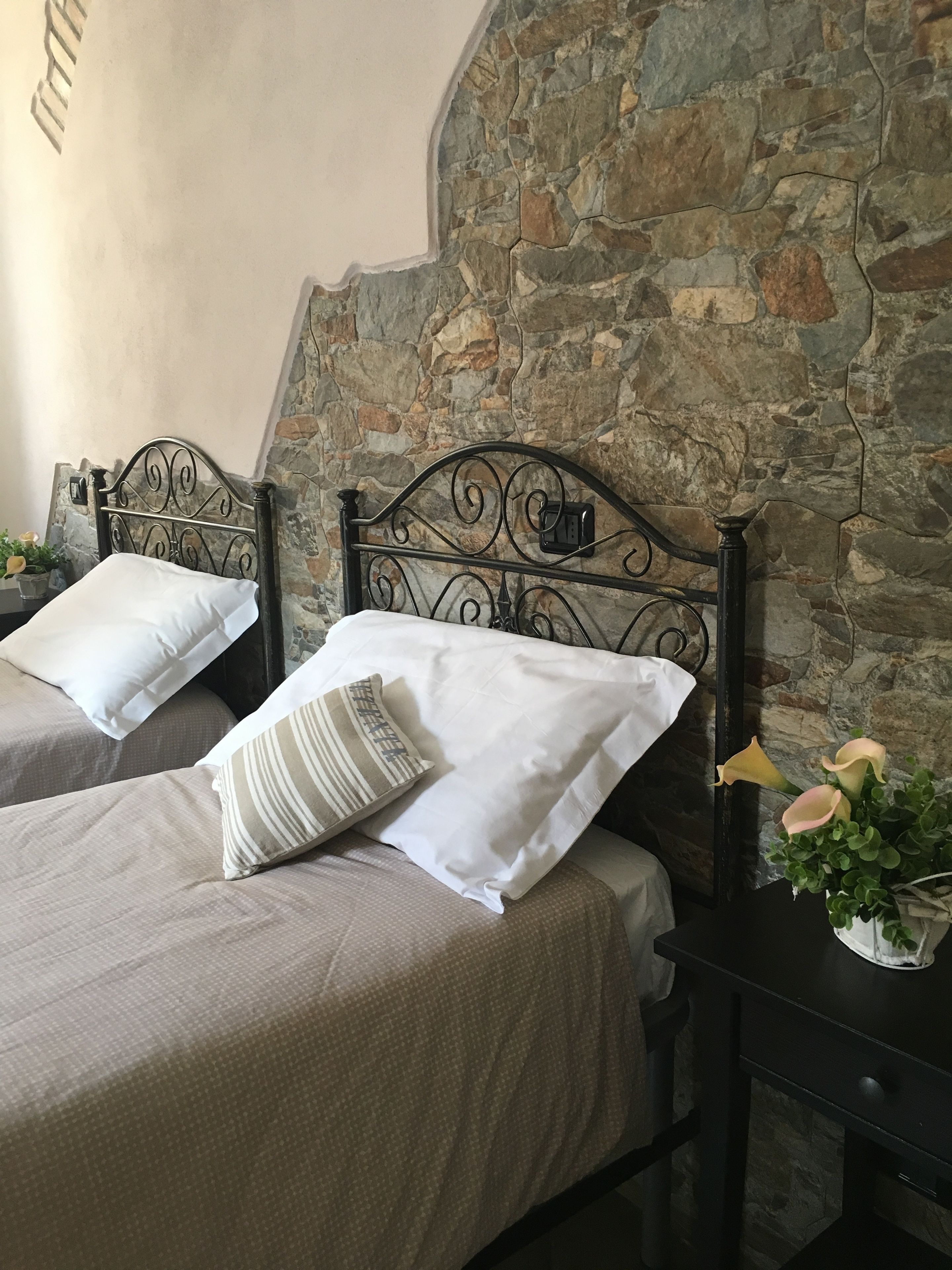 Due letti singoli con testiere in metallo addossati a un muro in pietra, un comodino con un vaso di fiori e un piccolo tavolino da notte con una lampada.