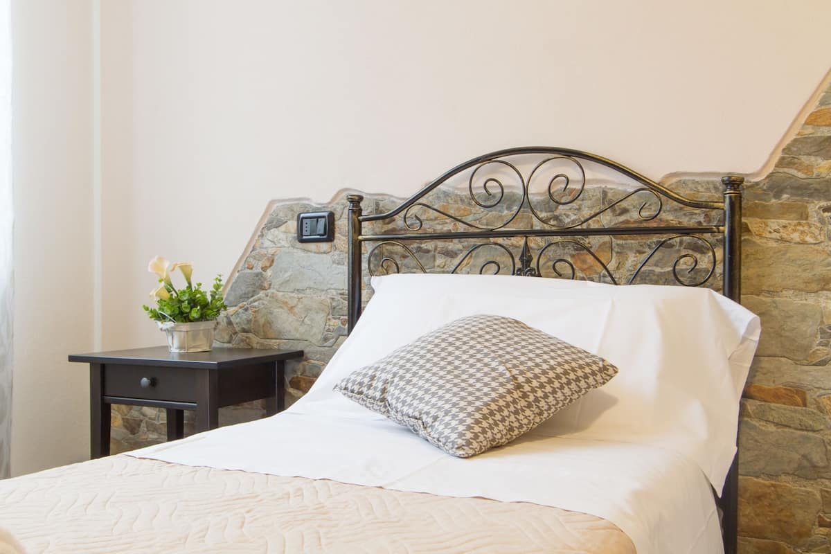 A neatly made bed with a patterned pillow, a small bedside table with a plant, and a stone wall behind the bed.