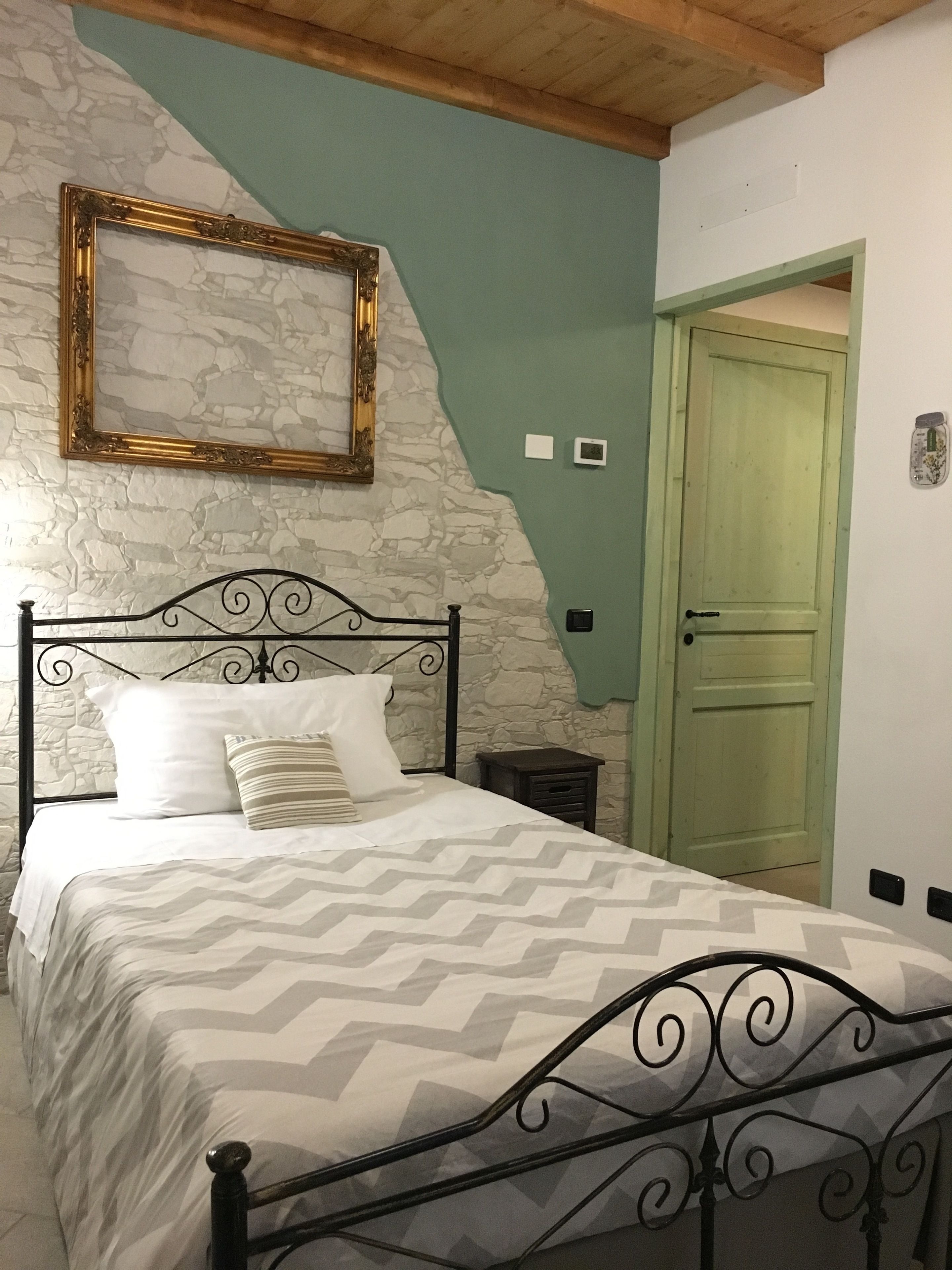 Un letto con struttura in metallo e copripiumino decorato, una parete in pietra, una porta verde e un quadro appeso al muro.