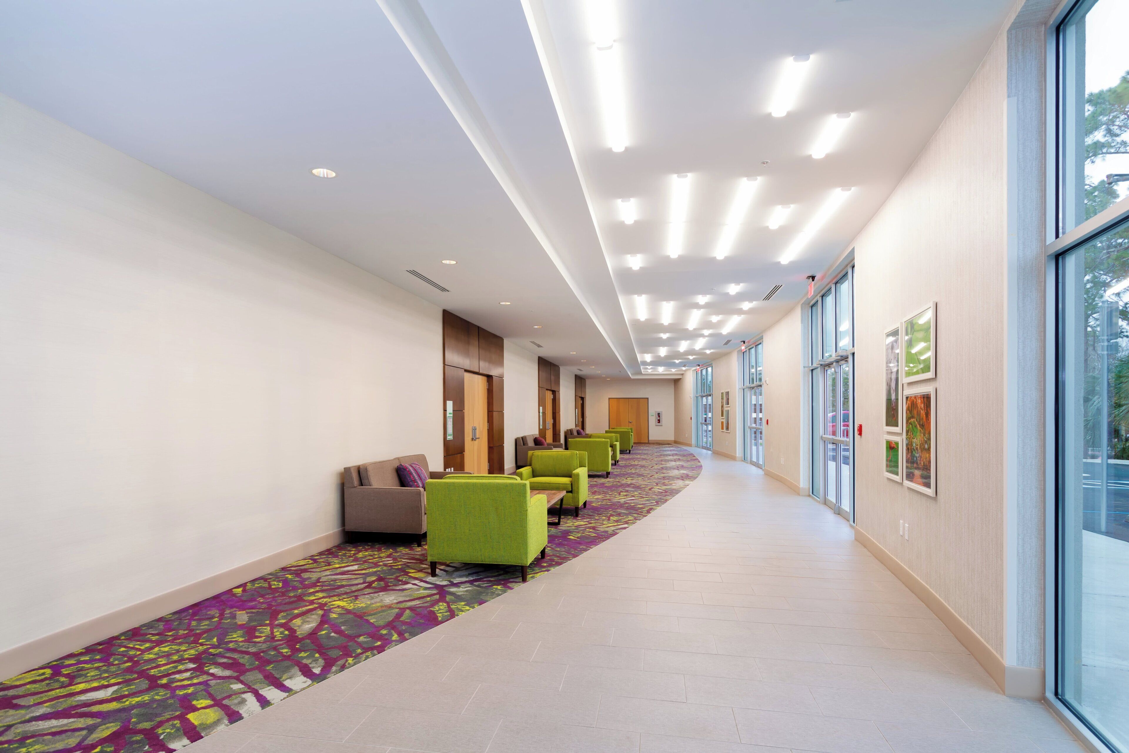 A hotel corridor with a row of green chairs, a patterned carpet, and large windows on one side.