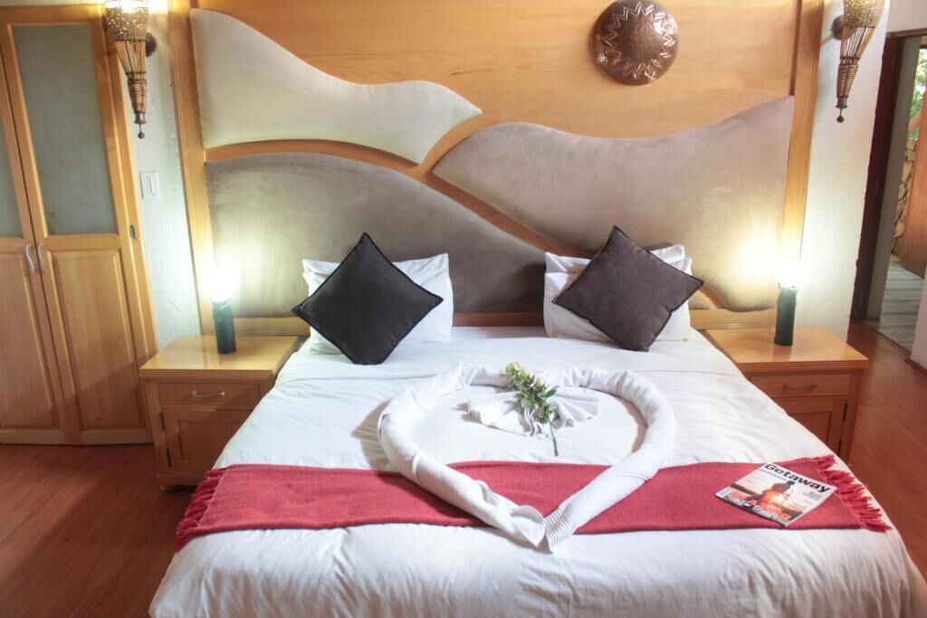 A neatly made bed with white linens, two black pillows, and a red throw blanket. There is a magazine on the bed and a small plant in a white vase. The room has wooden floors and a wooden headboard with a decorative light fixture above it.