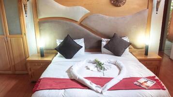 Una cama bien hecha con sábanas blancas, dos almohadas negras y una manta roja. Hay una revista sobre la cama y una planta pequeña en un jarrón blanco. La habitación tiene pisos de madera y un cabecero de madera con una lámpara decorativa encima.