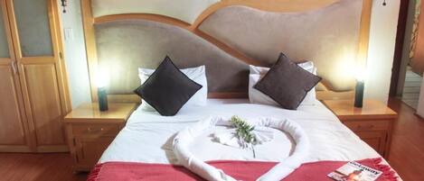 A neatly made bed with white linens, two black pillows, and a red throw blanket. There is a magazine on the bed and a small plant in a white vase. The room has wooden floors and a wooden headboard with a decorative light fixture above it.