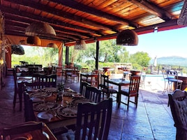 Un área de comedor al aire libre con mesas y sillas de madera, una piscina y vistas a zonas verdes y montañas.
