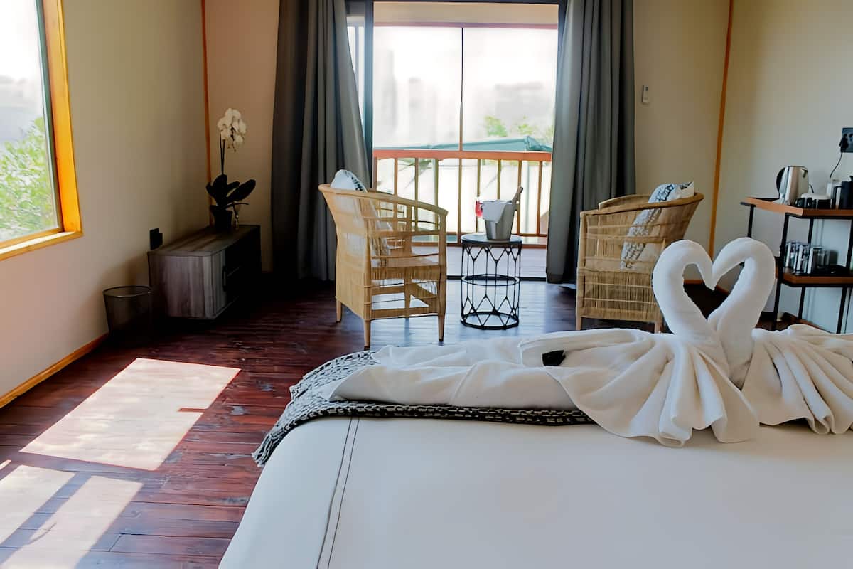 Ein Hotelzimmer mit einem Bett, einem kleinen Tisch mit einer Flasche, zwei Korbstühlen, einem Balkon mit Blick und einem Blumentopf.