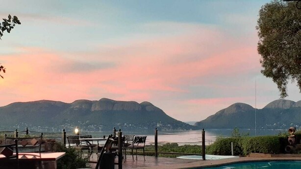 Uma área à beira da piscina com cadeiras e mesas, com vista para um lago e montanhas.
