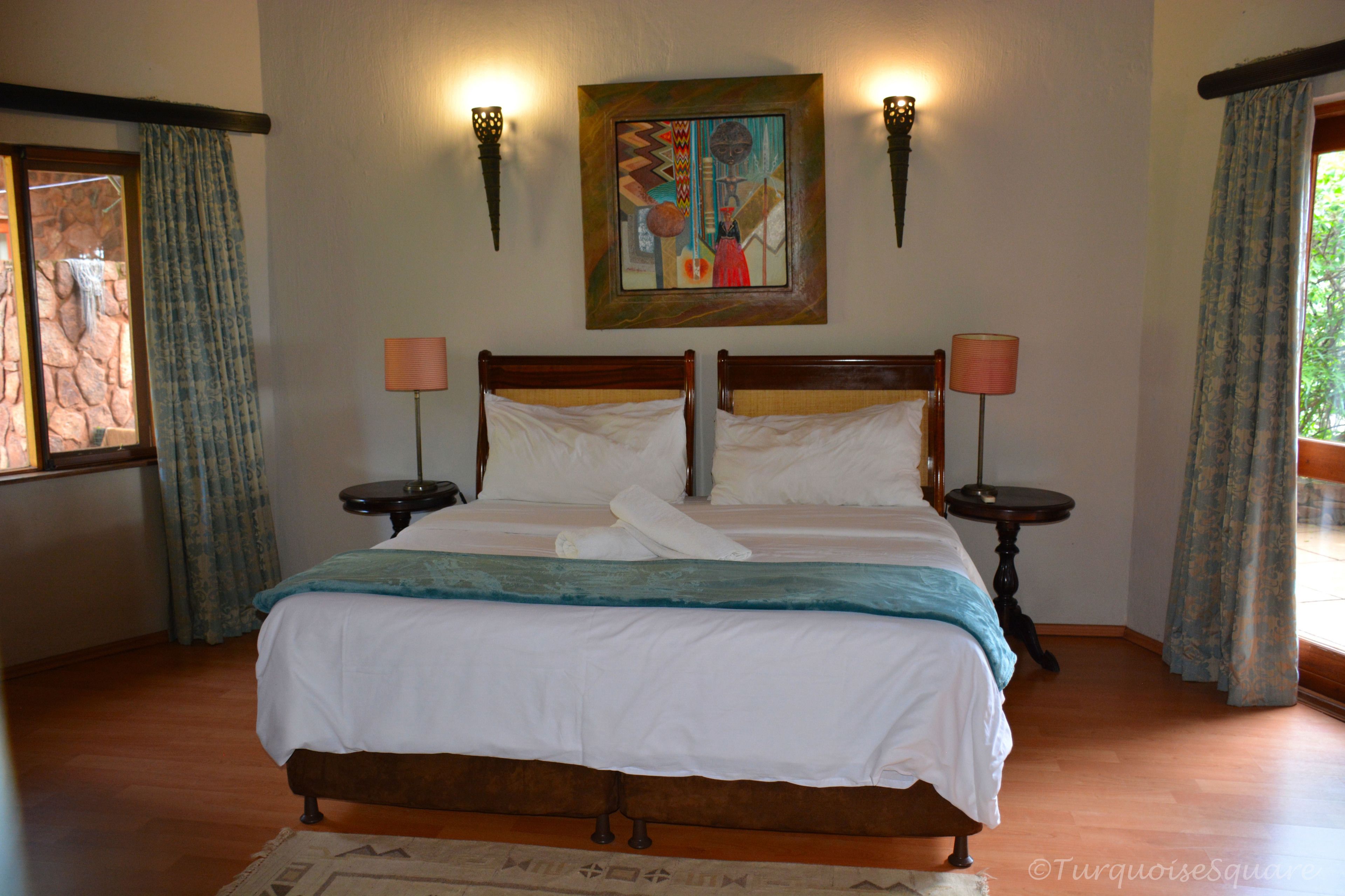 A double bed with white linens, two bedside tables with lamps, a painting above the bed, and a window with curtains.