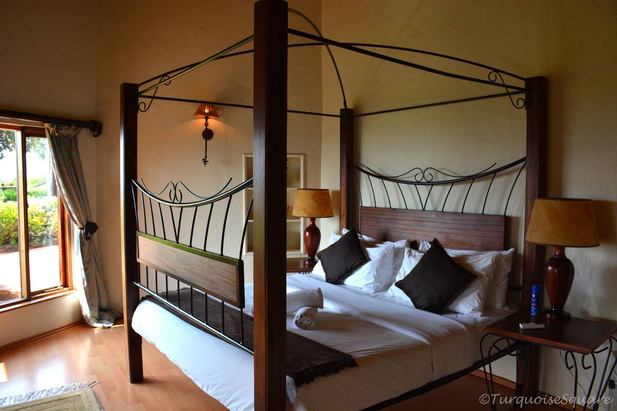 Ein Bett mit Baldachin, das ein hölzernes Kopfteil, weiße Bettwäsche und viele Kissen hat. Auf den Nachttischen stehen jeweils zwei Nachttischlampen.