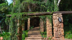 Uma escadaria de pedra leva a uma entrada coberta, rodeada por vegetação exuberante e um vigário de madeira.