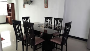Een eetruimte met een glazen tafel, omringd door zes stoelen, een televisie aan de muur en een boekenkast op de achtergrond.