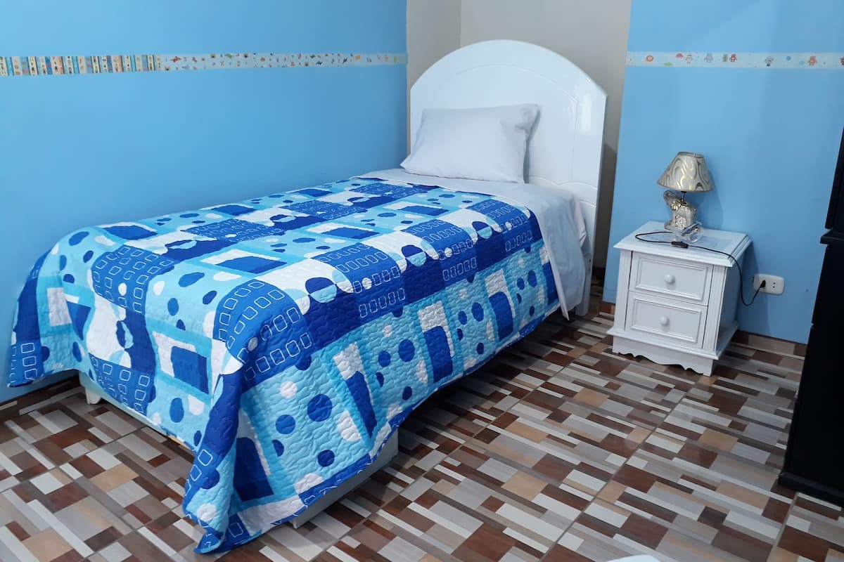 A bedroom with a bed covered in a blue and white patterned bedspread, a white headboard, a bedside table with a lamp, and a tiled floor.