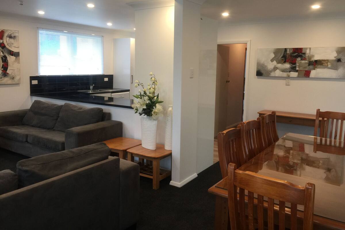 A modern living room with a grey sofa, a wooden dining table, and a kitchen in the background.