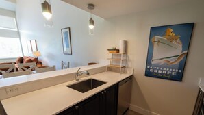 Una cocina moderna con un lavamanos, lavavajillas y luces colgantes. Se observa un cuadro en la pared y un cartel de un velero.