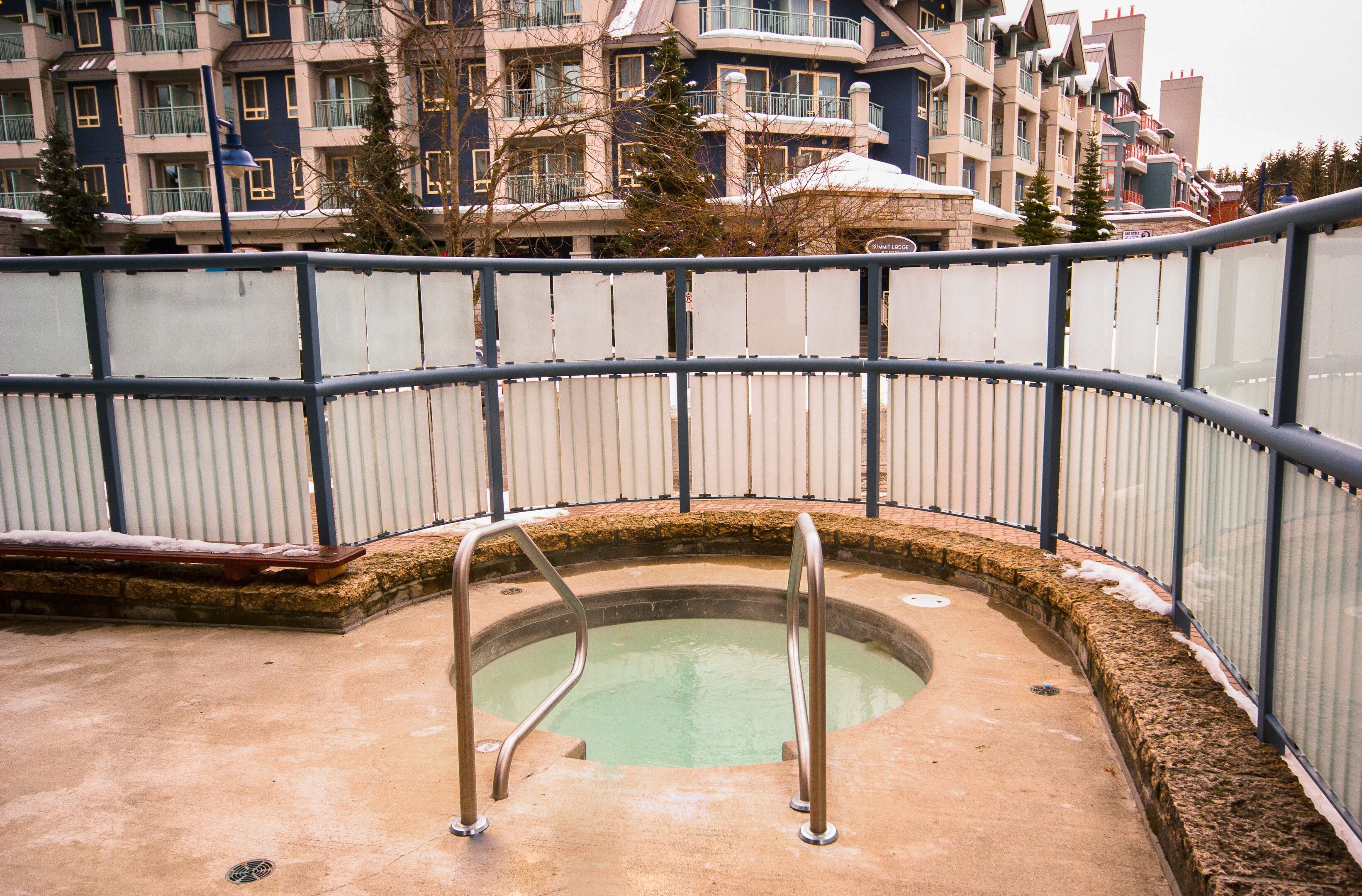 Zona de piscina con barandilla metálica, jacuzzi y vista a un edificio de varios pisos al fondo.