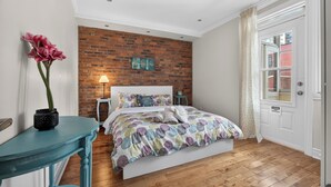 A bedroom with a brick wall, a bed with a colorful circular-patterned bedspread, a nightstand, a lamp, and a potted plant.