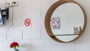 A round mirror on a white brick wall, a small table with chairs, a vase with red flowers, and a no-smoking sign.