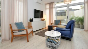 A modern living room with a sofa, armchair, and a round coffee table. There is a TV mounted on the wall, a small side table, and a potted plant. - Our flat is a great base to explore everything that South Australia offers... (West Beach)