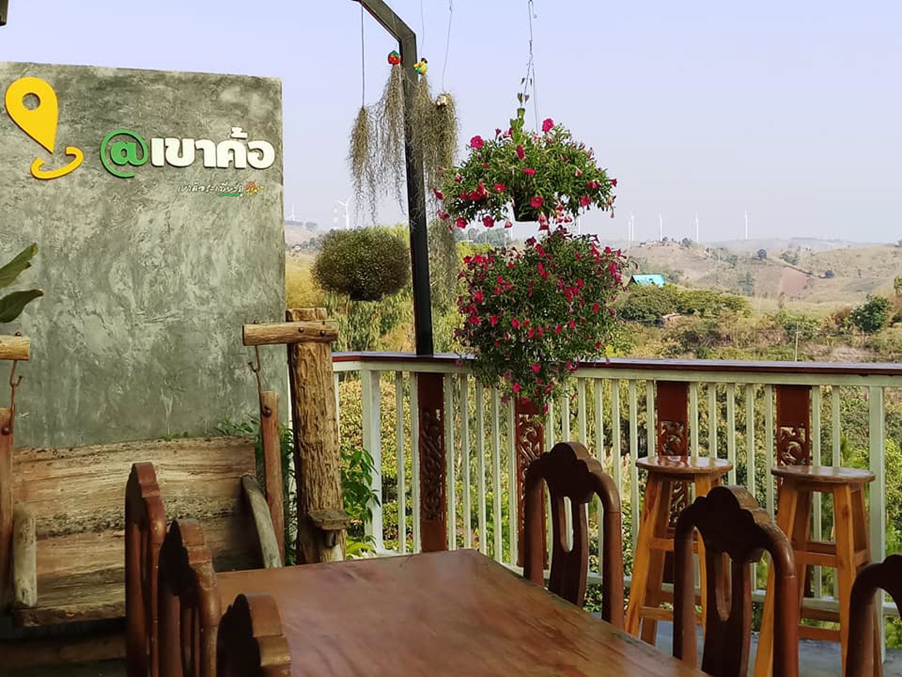 Una terraza de madera con una mesa y sillas, una cesta colgante de flores y vistas a parques eólicos a lo lejos.