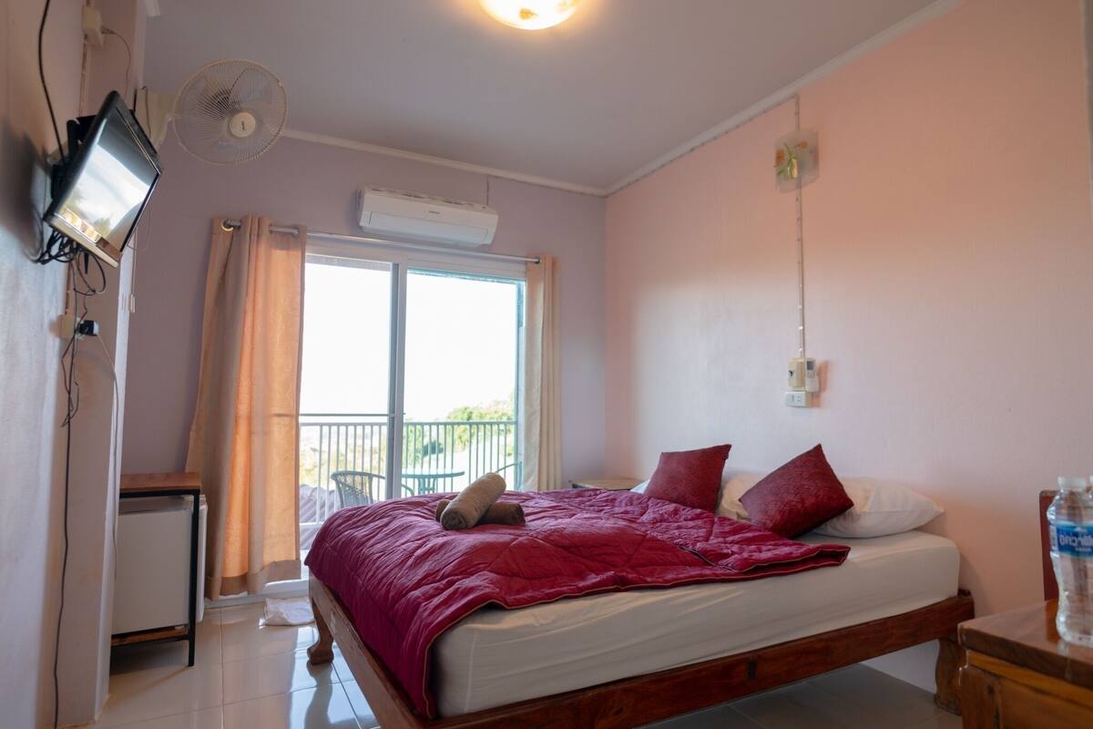 Ein Schlafzimmer mit Bett, Fernseher, Ventilator, Fenster mit Vorhängen und einem Balkon.