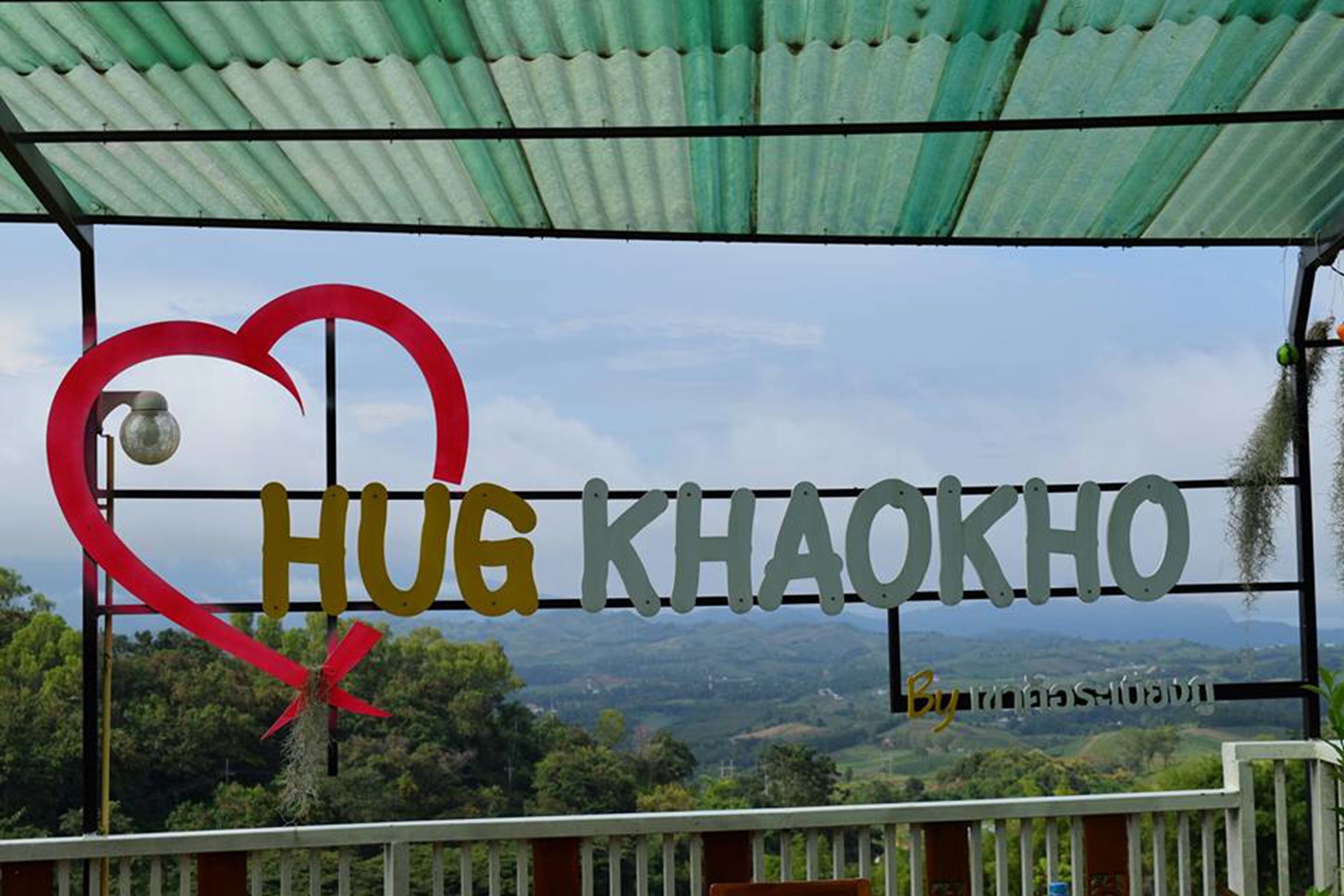 Un letrero con 'HUG KHAOKHO' y un símbolo de corazón, con vistas a un paisaje montañoso de fondo.