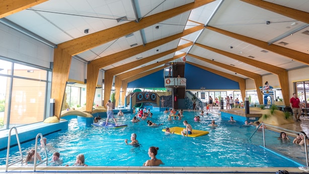 An indoor swimming pool with wooden beams, a lifeguard tower, and a variety of people swimming and playing.