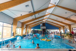 An indoor swimming pool with wooden beams, a lifeguard tower, and a variety of people swimming and playing.