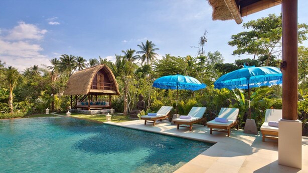A clear pool with lounge chairs and umbrellas, surrounded by tropical vegetation and a thatched-roof structure.