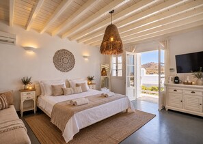 A bedroom with a large bed, bedside tables, a hanging light fixture, a television, and a view of the outdoors. - SEA & SUN A Quality Brand Certified Villa 150 meters from Agia Anna Beach (MYKONOS)