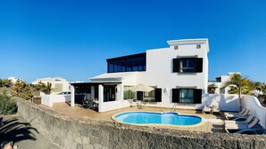 A modern two-story house with a swimming pool, outdoor seating, and a stone retaining wall. - Faro Park, Luxury Villa (VV), heated pool, Sea views,500m shops/bars/restaurants (Playa Blanca)