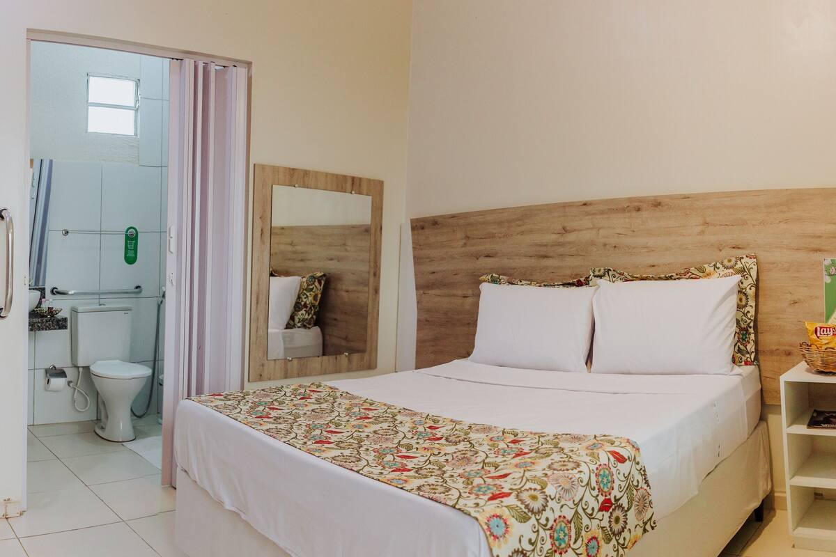 A hotel room with a bed, a desk with a plant, a TV, a bathroom, and a wooden headboard.
