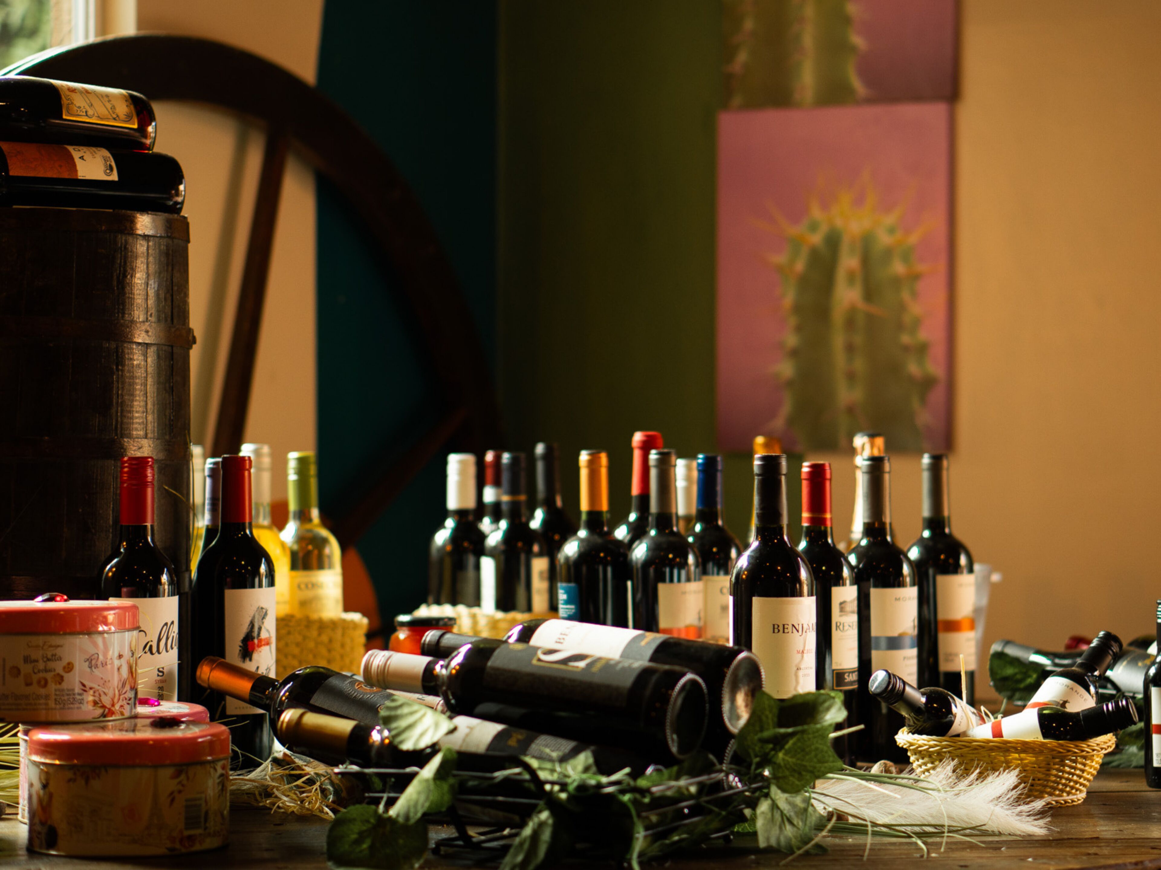 A variety of wine bottles and containers on a table, with a large wooden barrel and some greenery.