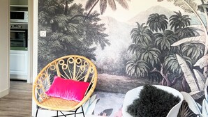 A room with a mural of tropical foliage, a wicker chair with a red cushion, and a rocking chair with a black cushion. - APARTMENT LA COLLINE 4 EPIS - Villas des Sables (Fréhel)