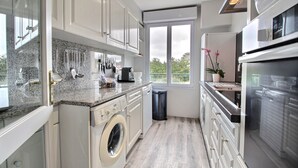 A modern kitchen with a washing machine, a coffee maker, and a view of the outdoors through a large window. - APARTMENT LA COLLINE 4 EPIS - Villas des Sables (Fréhel)
