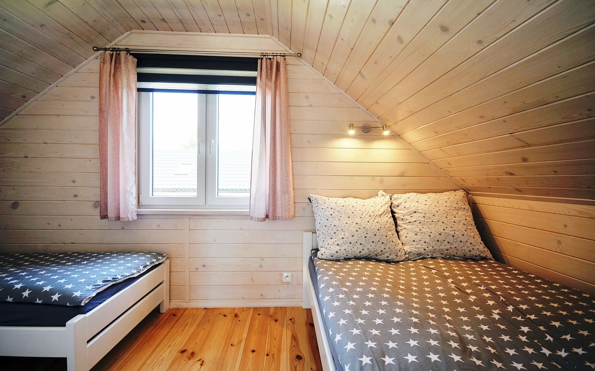 Ein kleiner, gemütlicher Raum mit schrägem Dach, einem fenster mit Vorhängen, einem Bett mit sternenmusternder Bettwäsche und einem kleinen Tisch.