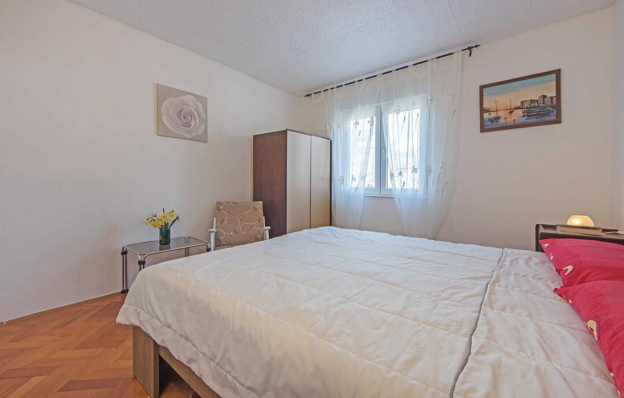 A bedroom with a bed, a chair, a wardrobe, a bedside table with a lamp, and a window with curtains.