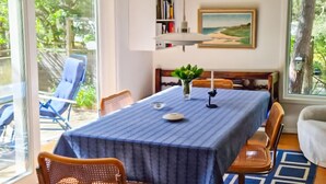 Ein Essbereich mit einem Tisch, der mit einer blauen Tischdecke bedeckt ist, umgeben von Korbstühlen. Ein Bücherregal mit Büchern und ein gerahmtes Gemälde an der Wand sind ebenfalls zu sehen.