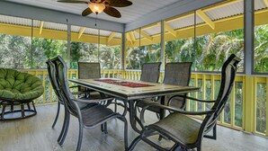 A covered outdoor dining area with a table and chairs, a ceiling fan, and large windows with a view of tropical vegetation. - Large comfy house big enough for 2 families next to ocean on Kaloli point (Keaau)