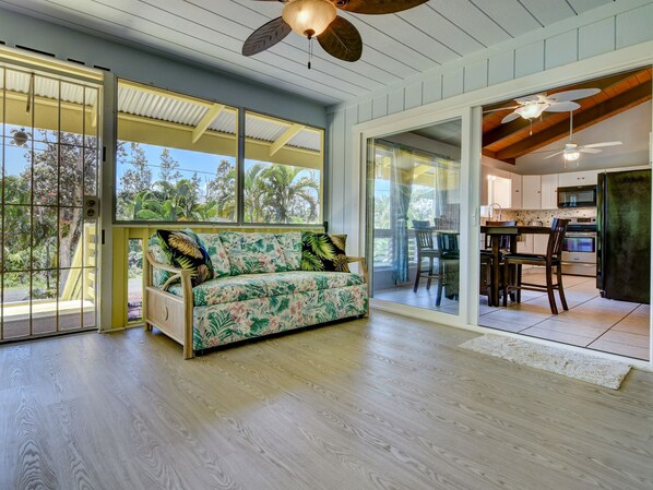 A spacious room with a wooden floor, a sofa with tropical prints, a ceiling fan, and a view of the outdoors.