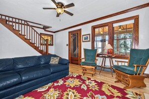 A living room with a blue leather sofa, two wooden chairs, a patterned rug, a ceiling fan, and a staircase.
