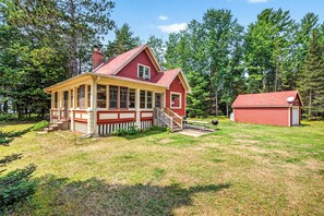 A red wooden cabin with a porch, a chimney, and a small shed in a wooded area. - Gorgeous lakefront cabin, wrap-around enclosed porch, dock! (St Germain)